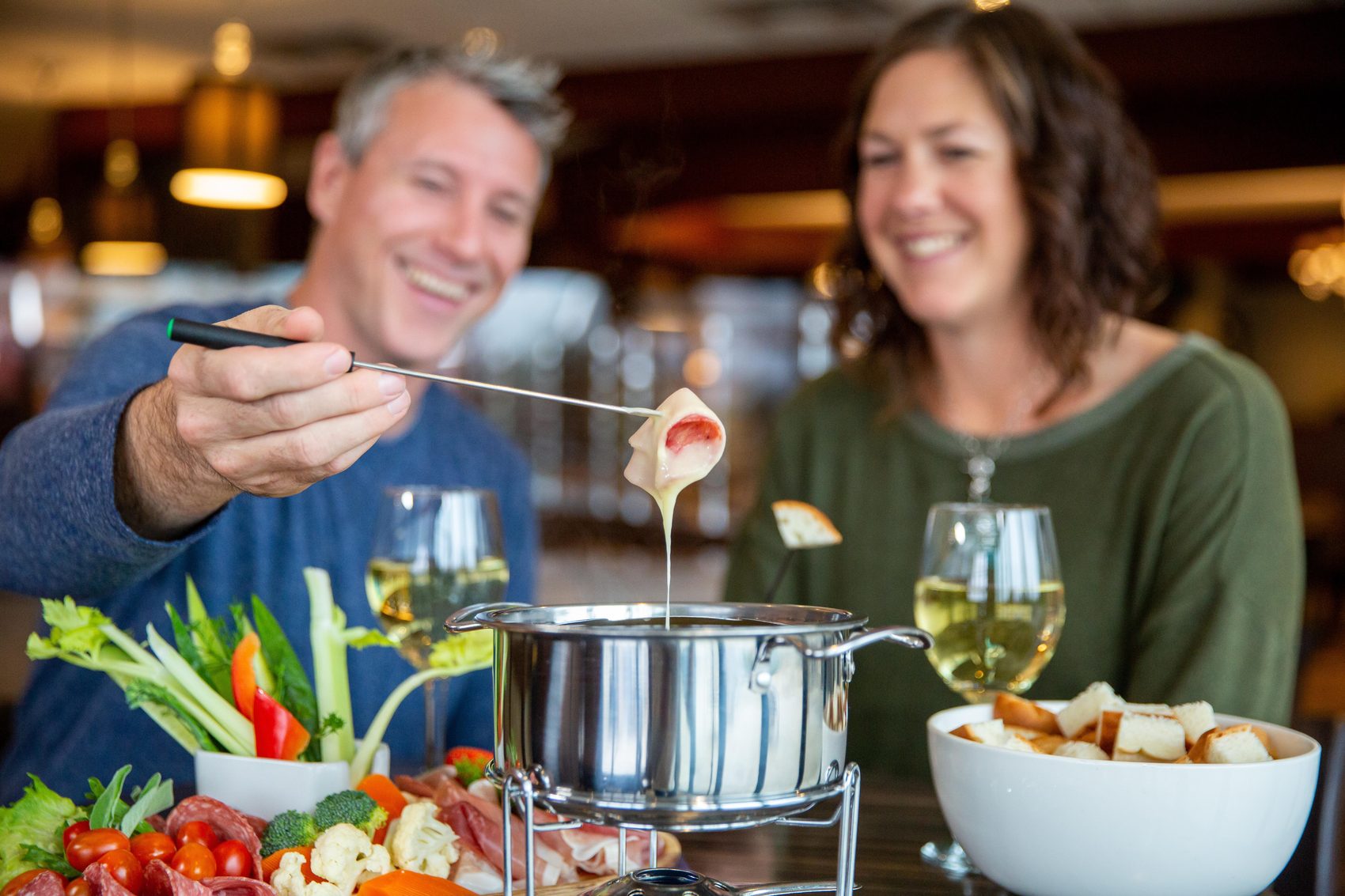 A couple share a fondue of vegetables and bread, with glasses of white wine, at a restaurant table