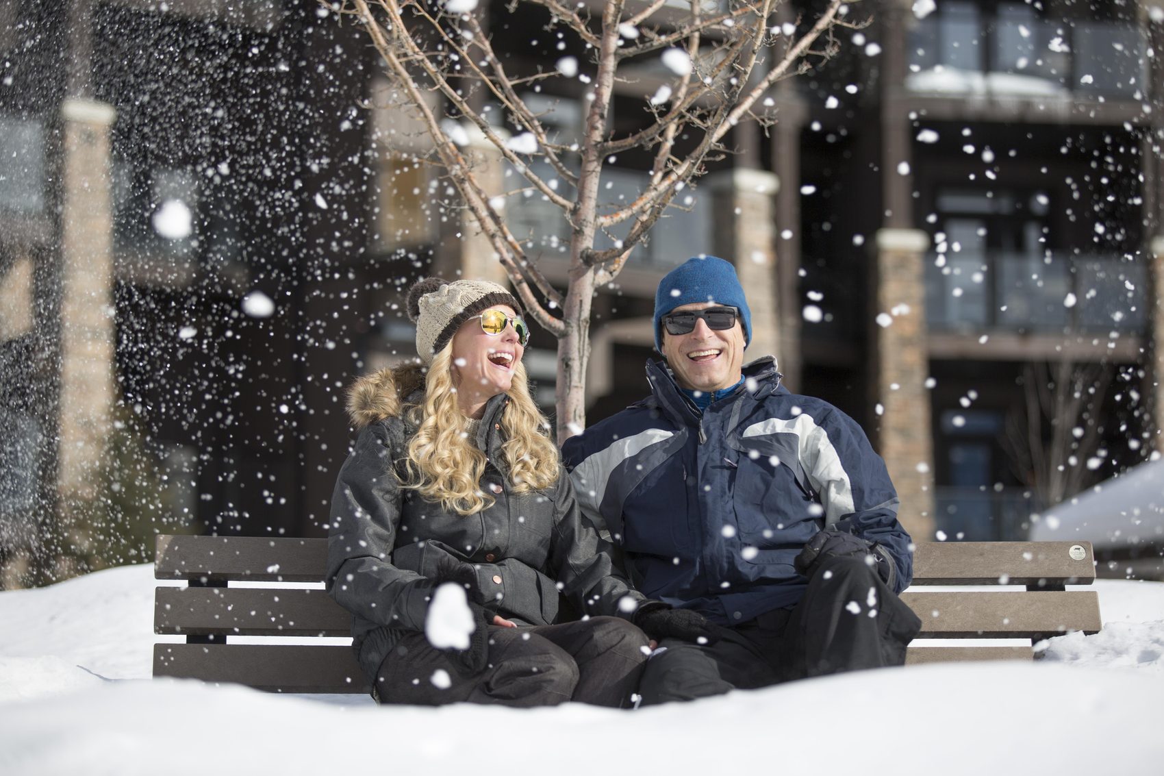 A couple in winter coats sit on a bench in falling snow