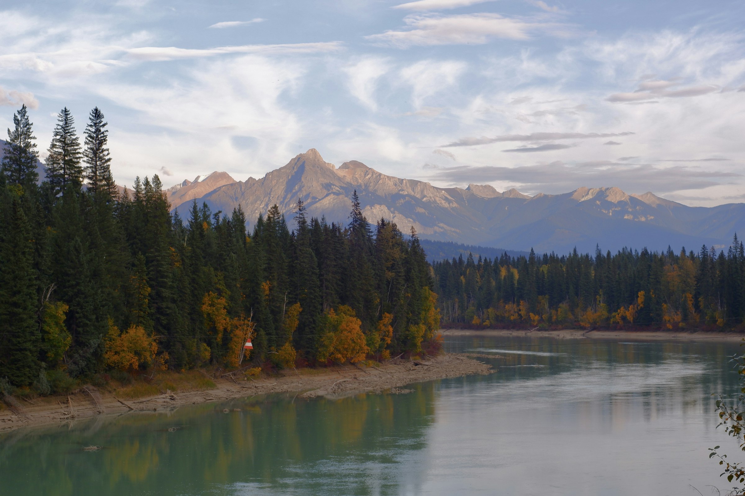 A calm river curves past conifers and autumn-coloured trees toward a mountain peak lit by late sun.